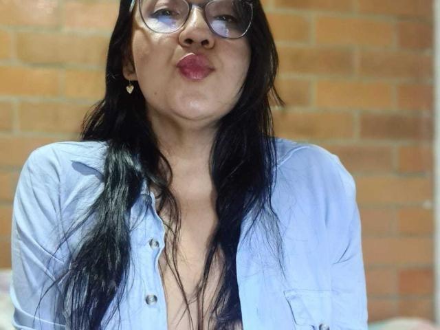 QueenSophiee with black hair, glasses, and pink lips, wearing an unbuttoned light blue shirt, exposing her cleavage, standing against a brick wall.