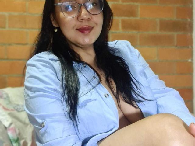 QueenSophiee, avec de longs cheveux noirs, des lunettes, une chemise bleu clair, et les seins apparents, assise avec un léger sourire, portant des boucles d'oreilles en forme de cœur, devant un mur de briques..