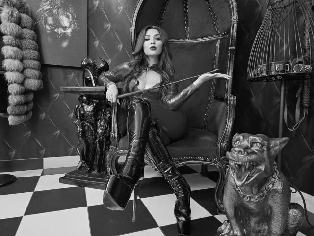 KayaOcean with long hair, dark lipstick, and black latex outfit, sitting in a vintage chair, holding a whip, wearing thigh-high boots, next to a grinning cat statue.