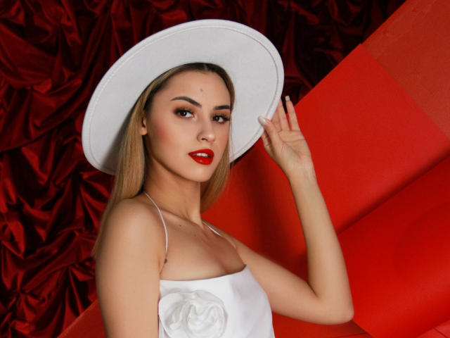 JasmineDay con cabello rubio, lápiz labial rojo, sombrero blanco y vestido blanco sin tirantes, sosteniendo el sombrero, mostrando senos pequeños, fondo rojo y pared roja, mirando a la cámara..