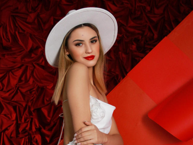 JasmineDay con cabello rubio, lápiz labial rojo, sombrero blanco y vestido blanco sin tirantes, con los brazos cruzados, sonriendo, mostrando sus pechos, sobre un fondo rojo y blanco..