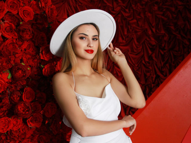 JasmineDay with blonde hair, red lipstick, white dress, and hat, holding it, standing against red roses, showing her tits, slim, attractive, sexy, confident, smiling.