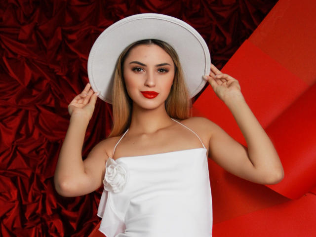 JasmineDay draagt rode lippenstift, een witte jurk en een bloem, en houdt een hoed met een brede rand vast. Ze heeft lang blond haar en kijkt naar de camera. Haar borsten zijn klein, ze is schattig..