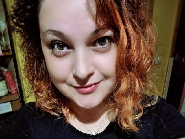 CurlyCherry has curly, reddish-orange hair, fair skin, and is smiling with pink lips, wearing a black shirt, and a silver necklace, with brown eyes looking at the camera.