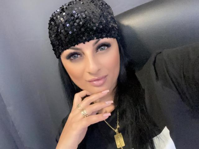 VixenSelena has black sequined cap, black top, long black hair, blue eyes, light pink lipstick, gold necklace, rings, and a confident smile, with her hand on her chin.