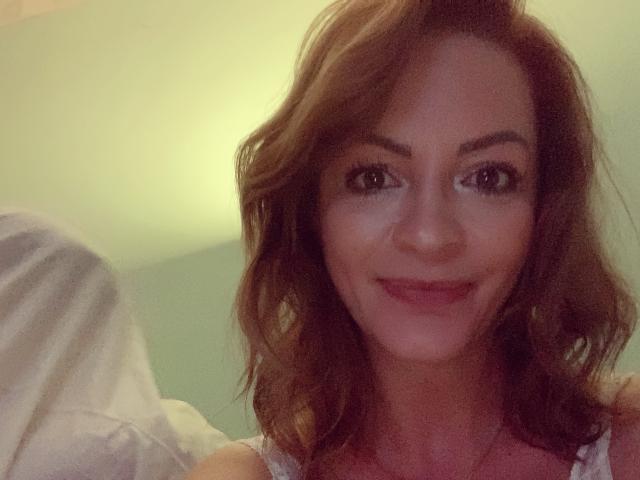 Cette enseignante, surnommée ThisTheacher69, a les cheveux bruns ondulés, les yeux marron et un sourire charmant avec des lèvres roses. Elle porte un haut rose, et la photo a une lumière douce et chaleureuse..