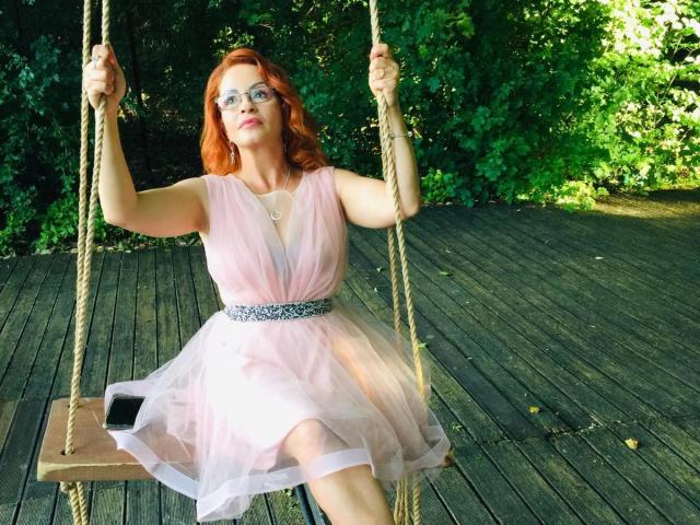 Cette enseignante, avec ses cheveux roux, ses lunettes et sa robe en tulle rose, est assise sur une balançoire en bois avec une corde, tenant la corde, un téléphone sur le siège, et du feuillage vert en arrière-plan. Elle porte une ceinture argentée..