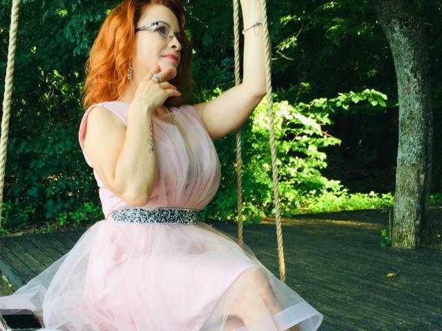 ThisTheacher69 with red hair, glasses, and a pink tulle dress, sitting on a wooden swing, holding rope, wearing a sparkly belt, looking to the side.
