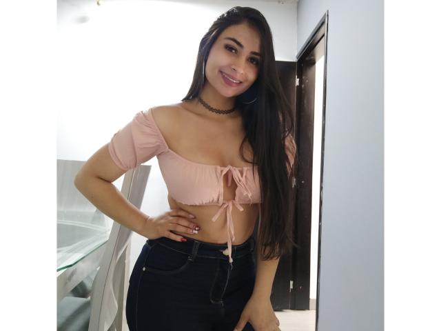 MiilaWx with long black hair, pink off-shoulder crop top, tied below her boobs, black high-waisted jeans, smiling, hand on hip, choker necklace, standing indoors.