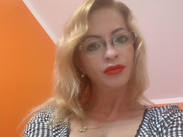 ThisTheacher69 a les cheveux blonds, porte des lunettes, du rouge à lèvres et une chemise à carreaux. Elle regarde l'appareil photo avec une expression sérieuse. Ses seins sont visibles sous la chemise..