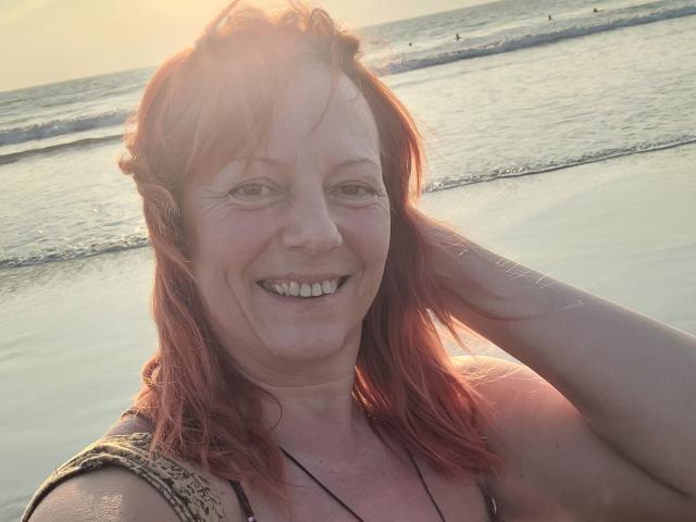 BarbaraBowie met rood haar, lachend, zonnig strand, hand in haar, draagt een top met een patroon, een ketting, lichte huid, zichtbare tanden, casual, warm licht, lichte lensflare, ontspannen..