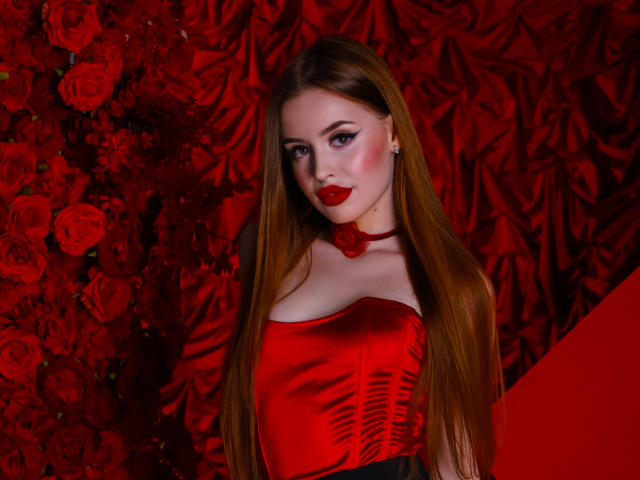 CherylFoxy with long red hair, red lipstick, and a red satin dress, showing her tits, standing against a red rose wall, looking at the camera with a slight smile.