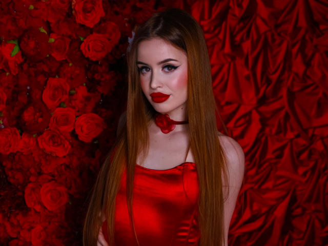 CherylFoxy, com cabelos ruivos longos, vestindo um vestido vermelho brilhante e um colar, batom vermelho e maquiagem nos olhos, em pé em frente a uma rosa vermelha e um fundo de cetim. Seios visíveis, fofa..