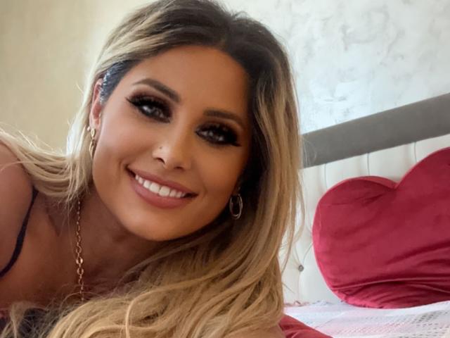Anca, una mujer con cabello rubio largo, ojos marrones y una sonrisa, lleva aretes de aro y un collar de oro, y se muestra en un selfie mostrando sus senos. En el fondo, hay un cojín con forma de corazón rojo..