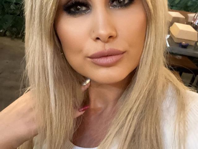 A AncaHotBodie tem cabelo loiro, olhos esfumados e lábios rosados, com uma mão tocando o pescoço, mostrando unhas vermelhas e um anel de prata, em uma selfie..