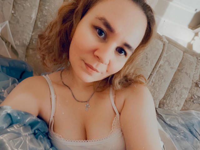 HotGirlHotLoves with curly brown hair, blue eyes, and cute smile, wearing a white lace bra, showing her tits, lying on a bed with a blue patterned blanket.