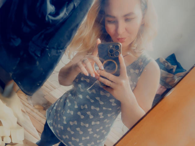Une jeune femme séduisante aux cheveux blonds, portant un haut gris à motifs, se prend en photo dans un miroir, tenant un téléphone avec une bague à l'annulaire, et regardant légèrement vers le bas..