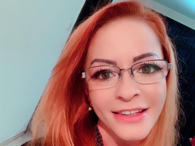 ThisTheacher69 has red hair, glasses, and is smiling with green eyes, wearing makeup and a black top, showing her nose and lips. She has a cute, sexy face.