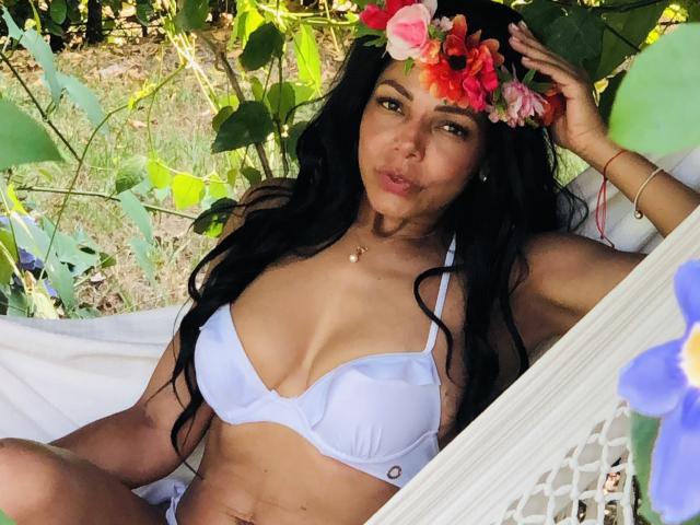VenusChaud with long black hair, wearing a white bra, flower crown, and gold necklace, sitting in a hammock, showing her tits, with green leaves and white cloth around.