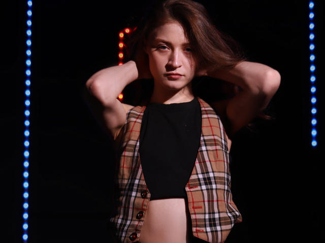 MiaBerry with arms raised, wearing a plaid sleeveless vest over a black crop top, revealing a flat stomach, and brown wavy hair, in a dark, lit-up setting.