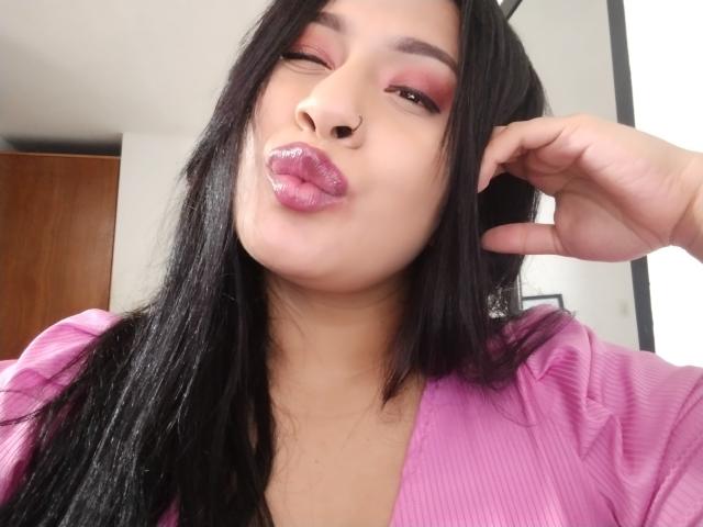 AlicTwenty with glossy pink lips, black hair, and a nose ring, winking with a hand in her hair, wearing a pink ribbed top. Her makeup is bold and sexy.