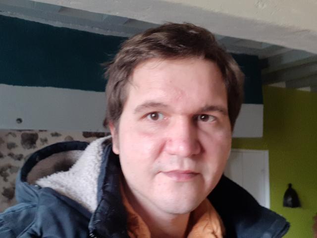 NectarineFad a un visage rond, des cheveux courts et bruns, et porte une veste matelassée bleu foncé avec un col en polaire blanc. Les yeux de NectarineFad sont marrons et son expression est neutre..