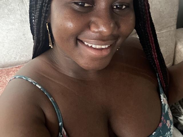Dilay has dark skin, braided hair with red highlights, small earrings, and is smiling at the camera, showing her tits in a floral dress, looking cute.