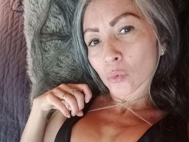 NicoletteShea with pouty lips, gray hair, brown eyes, black top, gold necklace, rings, and a chain, lying down, hand near face. Boobs visible, looking at camera.