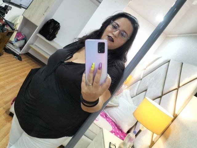 SkayLaraWalker, avec ses cheveux foncés, ses lunettes et son rouge à lèvres, tenant un téléphone, prenant un selfie devant un miroir, portant un haut noir, un pantalon blanc et des ongles jaunes, dans une pièce très éclairée..