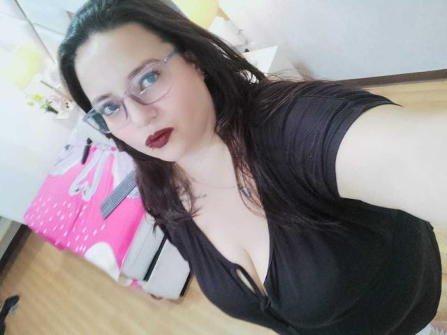 SkayLaraWalker, avec des lunettes, du rouge à lèvres rouge foncé et un haut noir, montrant de grandes seins dans un selfie, avec des cheveux bruns et une peau pâle, debout dans une pièce avec un lit rose..