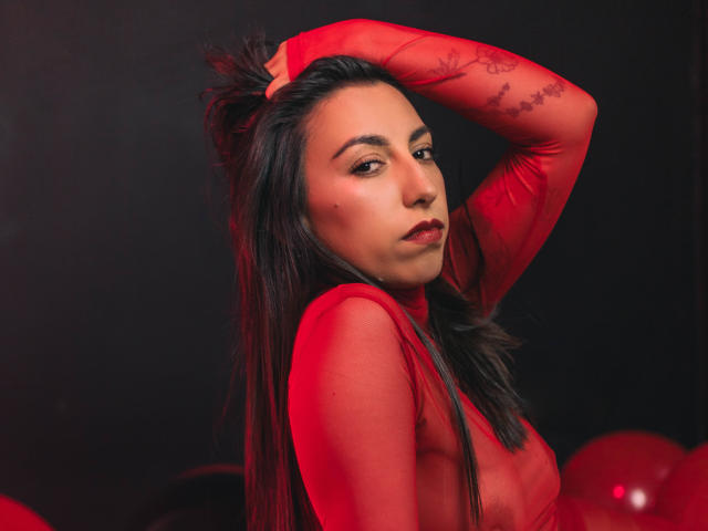 KeitlyBlue con cabello oscuro, blusa roja transparente de manga larga, y brazo levantado, mirando a la cámara, rostro iluminado en rojo, maquillaje sutil, pequeña peca en la mejilla, globos rojos en el fondo..
