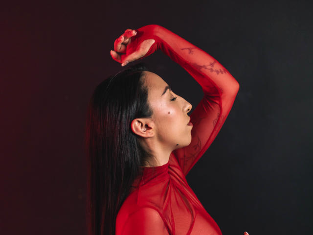 KeitlyBlue mit langen, schwarzen Haaren, einem roten, transparenten Kleid und erhobenen Armen, die Tattoos an ihren Armen und an der Seite zeigen, mit einem Muster aus Sommersprossen auf ihrer Wange. Sie blickt nach oben..