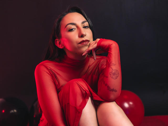 KeitlyBlue mit langen, schwarzen Haaren, trägt ein durchsichtiges, rotes Kleid und sitzt mit angezogenen Knien, die Hand am Kinn. Im Hintergrund rotes Licht, ein Tattoo am Unterarm, ein verführerischer Ausdruck, rote Ballons im Hintergrund..