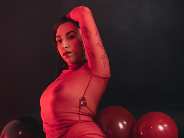 KeitlyBlue em um vestido vermelho transparente, com os braços levantados, mostrando seios pequenos e tatuagens, com balões vermelhos ao fundo, iluminados em vermelho. Ela tem cabelo escuro e olhos sedutores..