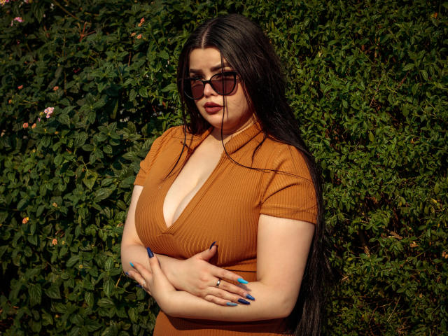 BrithneyLee with long black hair, brown ribbed top, cleavage, crossed arms, blue nails, sunglasses, and a ring, standing against green foliage.