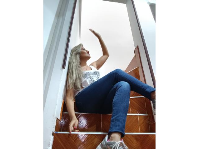 ValeriaHunter69 sits on wooden stairs with arms raised, wearing a white tank top, blue jeans, and gray sneakers, blonde hair flowing, eyes looking upward, legs crossed.