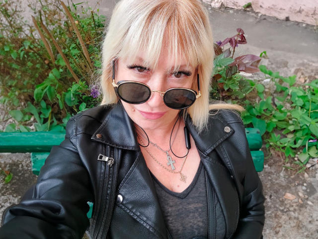 JessieFresh with blonde hair, black sunglasses, and black leather jacket, smiling at the camera, wearing a gray top and multiple necklaces, sitting on a green bench.