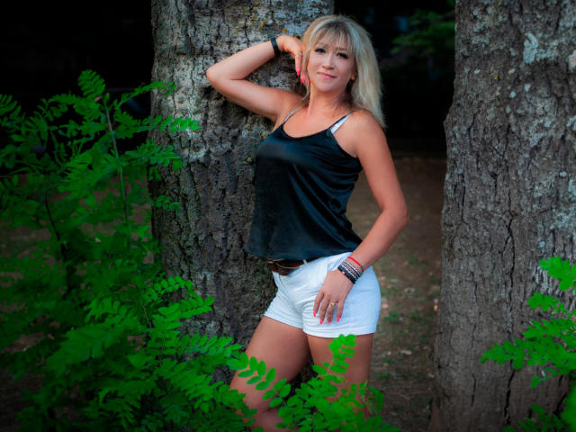 JessieFresh with blonde hair, black tank top, and white shorts, leaning against a tree, hand on head, smiling, wearing bracelets, green foliage in front, standing between tree trunks.