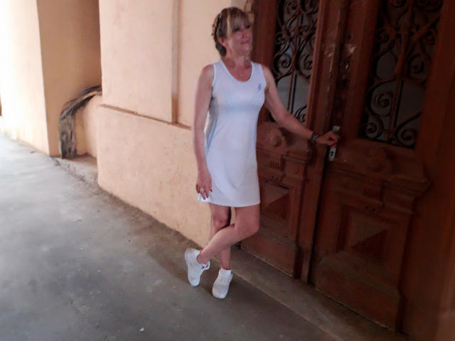 JessieFresh stands near a wooden door, wearing a white sleeveless dress, white sneakers, and holding a water bottle. She has short blonde hair and a small ass.