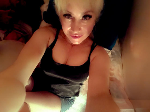 JessieFresh, avec ses cheveux blonds, portant une robe en satin noir, montrant de grandes seins, assise sur un lit, prenant un selfie avec un sourire sexy, les jambes croisées..