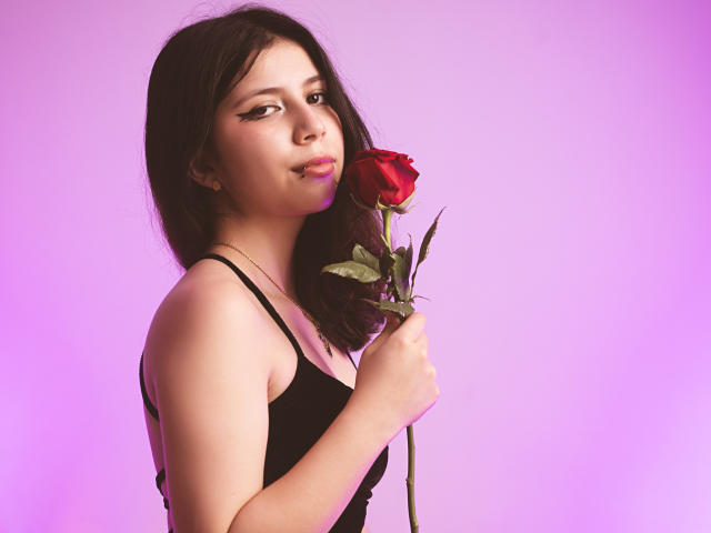 EmilyMalhore holding a red rose with green leaves, wearing a black tank top, purple background, looking at the camera with a slight smile, medium-length brown hair.