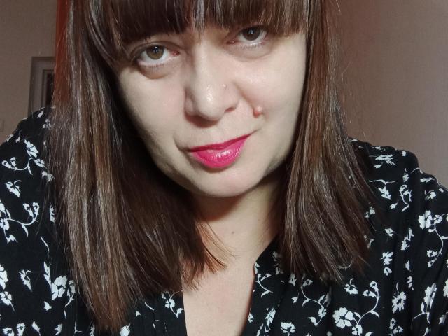 A PerkyBoobsMature tem olhos castanhos, batom rosa e cabelo castanho reto com franja. Ela está vestindo uma camisa preta com estampas florais, sorrindo levemente e tem uma pequena perfuração no nariz à esquerda..