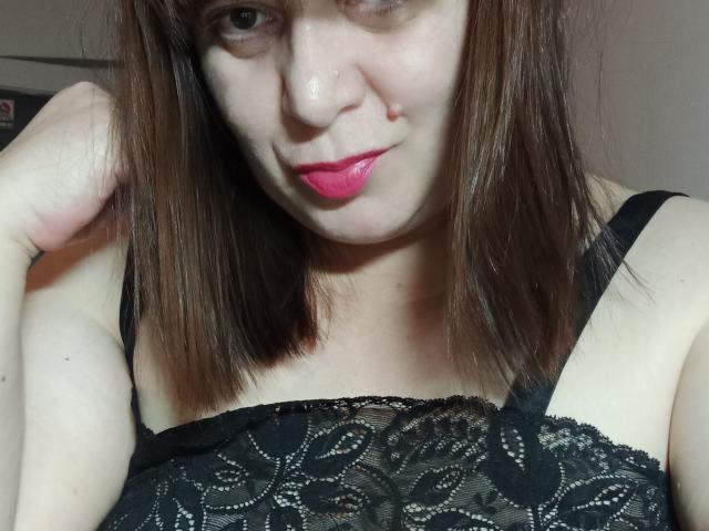 PerkyBoobsMature mit mittelbraunen Haaren, rosa Lippenstift und einem schwarzen Spitzen-BH, zeigt ein leichtes Lächeln, mit der Hand in der Nähe ihres Kopfes. Die Brüste sind durch die Spitze sichtbar..