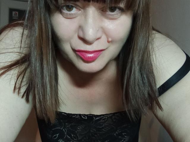 PerkyBoobsMature con cabello castaño, flequillo, lápiz labial rosa, top de encaje negro y correa negra, inclinada hacia adelante, sonriendo, tocándose la nariz, piel pálida, busto medio, escote ligeramente visible..