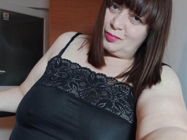 PerkyBoobsMature met bruin haar, roze lippenstift, een zwarte kanten top, waarbij haar grote borsten zichtbaar zijn, lichte huid en een lichte glimlach, die een selfie maakt in een huiselijke omgeving..