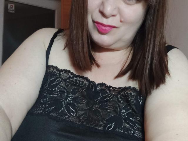 PerkyBoobsMature com cabelo castanho médio, batom rosa, blusa de renda preta, mostrando o peito, os braços e um leve sorriso. Os seios dela estão cobertos por renda. Ela está tirando uma selfie..