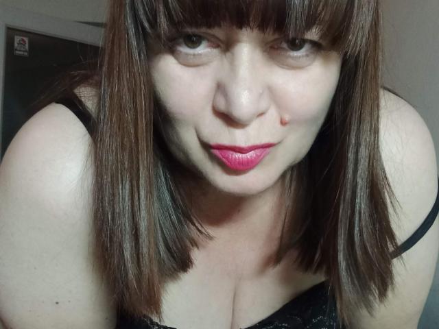 PerkyBoobsMature met bruin, steil haar, roze lippenstift en een schattige neus, leunt voorover en laat haar decolleté zien in zwart kant, heeft een kleine rode vlek op haar rechterwang..