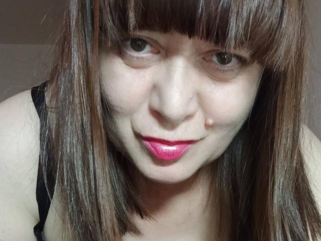 PerkyBoobsMature com cabelo castanho liso, franja, batom rosa, sorrindo levemente, top de alças pretas, pele clara, pequena marca de beleza na bochecha direita, olhos verdes, selfie de perto..