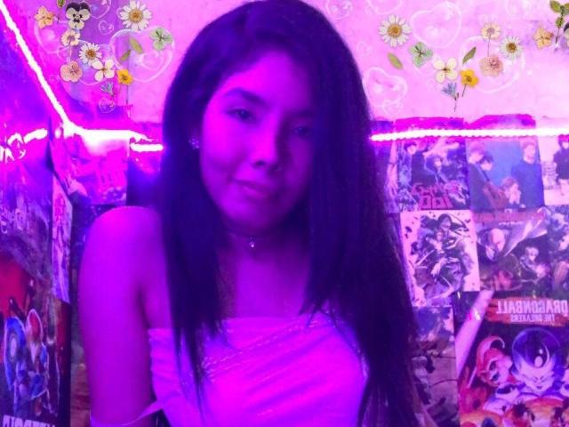 MileyWatson with long dark hair, purple-lit face, smiling, wearing a white top, in a room filled with anime posters. Her ass is not visible.