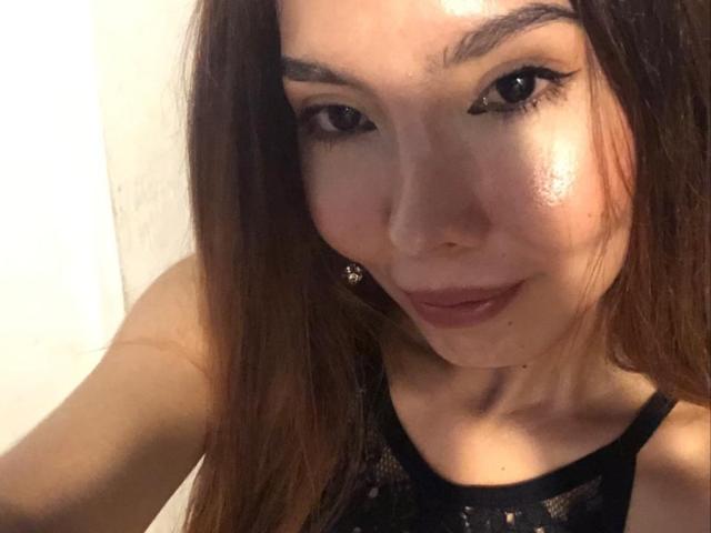MileyWatson has long brown hair, black eyeliner, and is wearing a black lace top. Her face is slightly shiny with a subtle smile. She has small earrings.